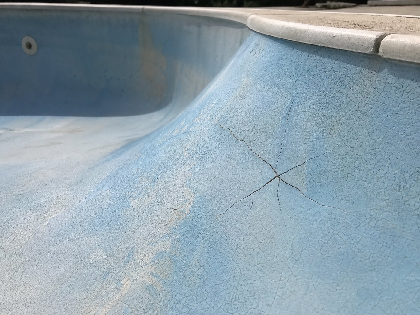 Spider web gel coat crazing and a star crack in the pale blue surface of a fibreglass swimming pool shell